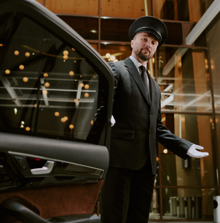 Caucasian middle aged man wearing uniform and cap opening car door with gloved hand, standing in modern building entrance, looking at camera, providing professional chauffeur service